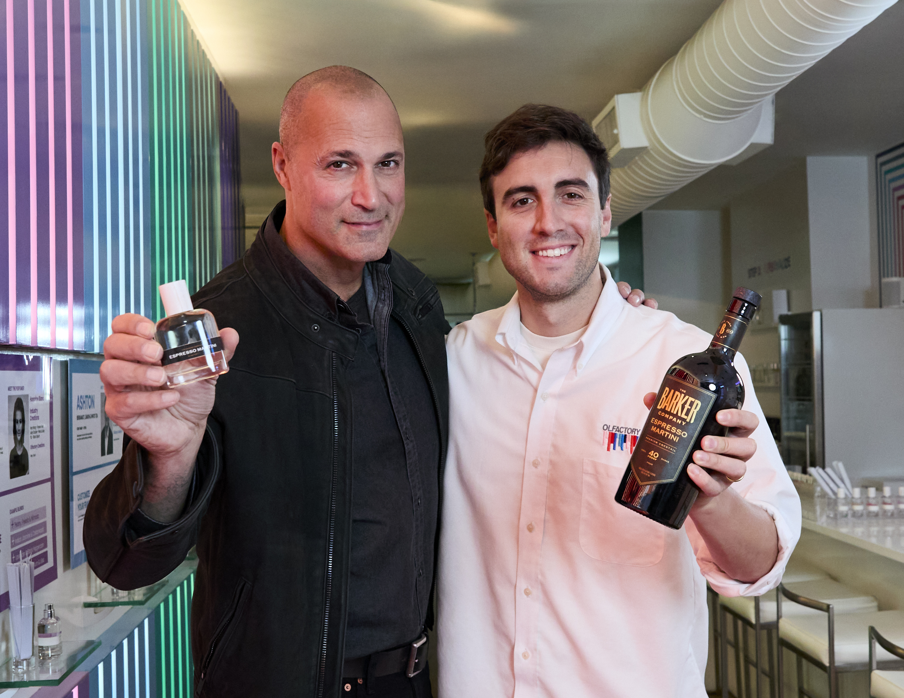 Two men smiling, holding a perfume bottle and a liquor bottle.