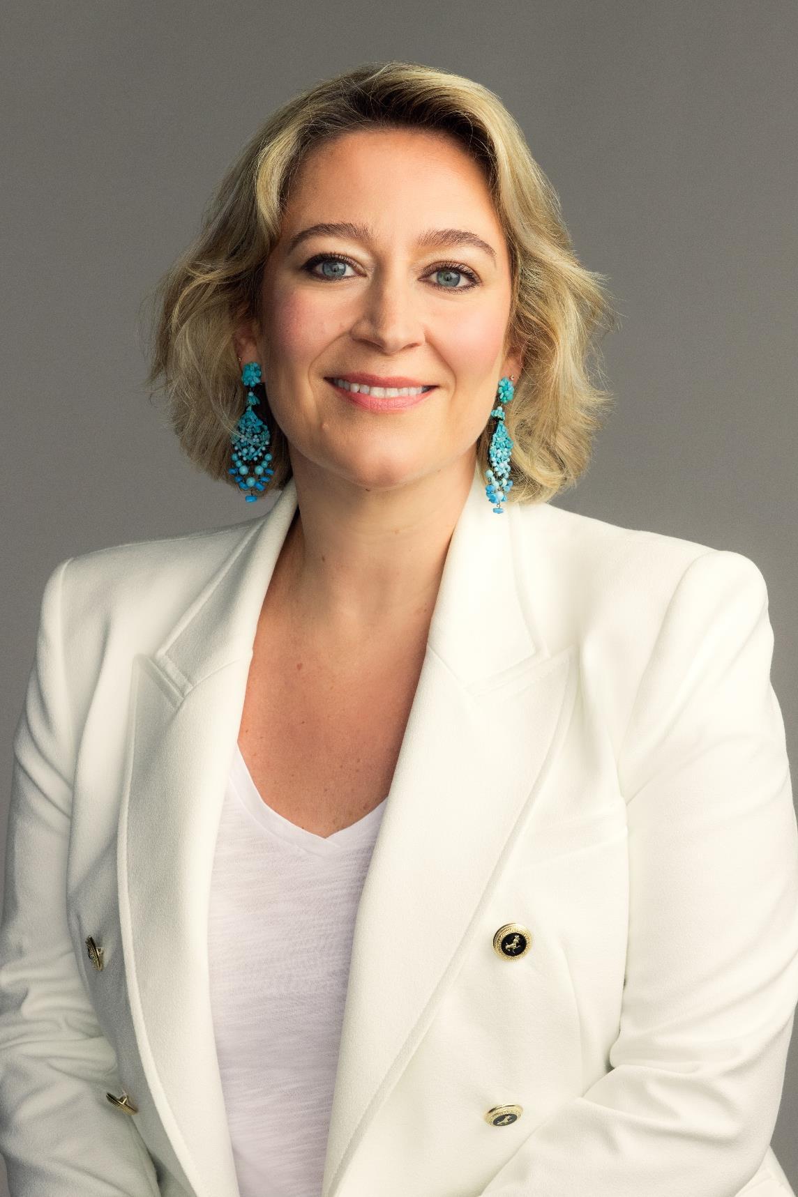 A smiling blonde woman in a white blazer and large turquoise earrings against a grey background.