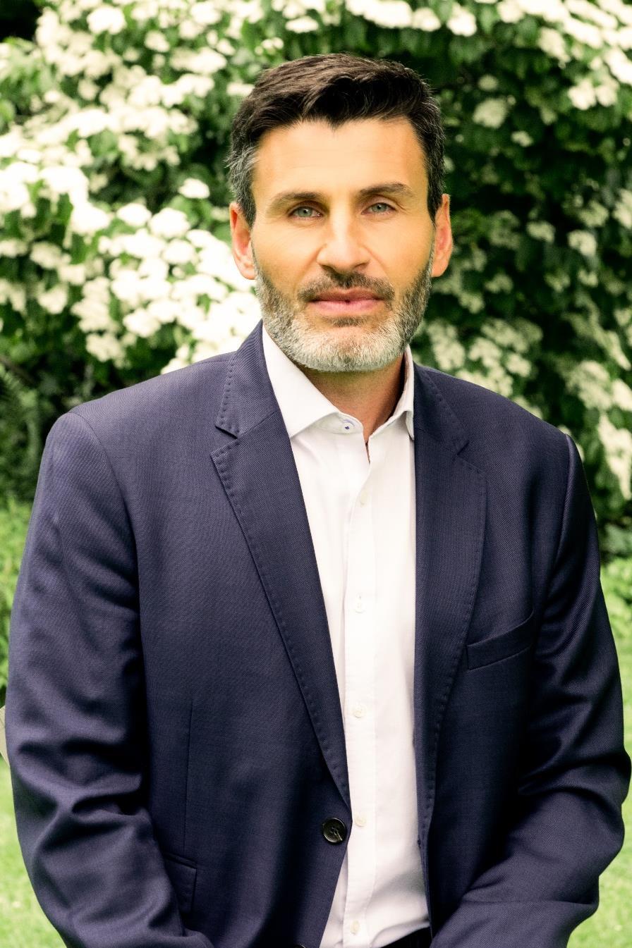 A man with a salt-and-pepper beard in a blue suit jacket standing in a garden.