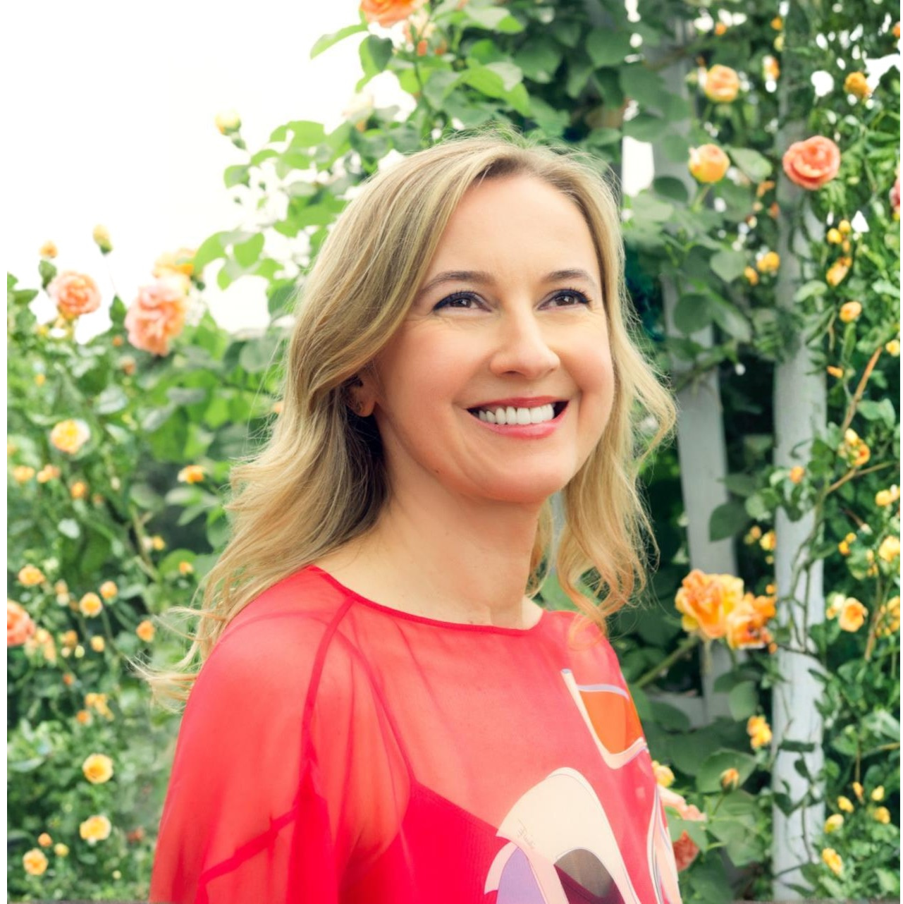 A smiling woman with blonde hair in a red top stands in front of a rose bush.