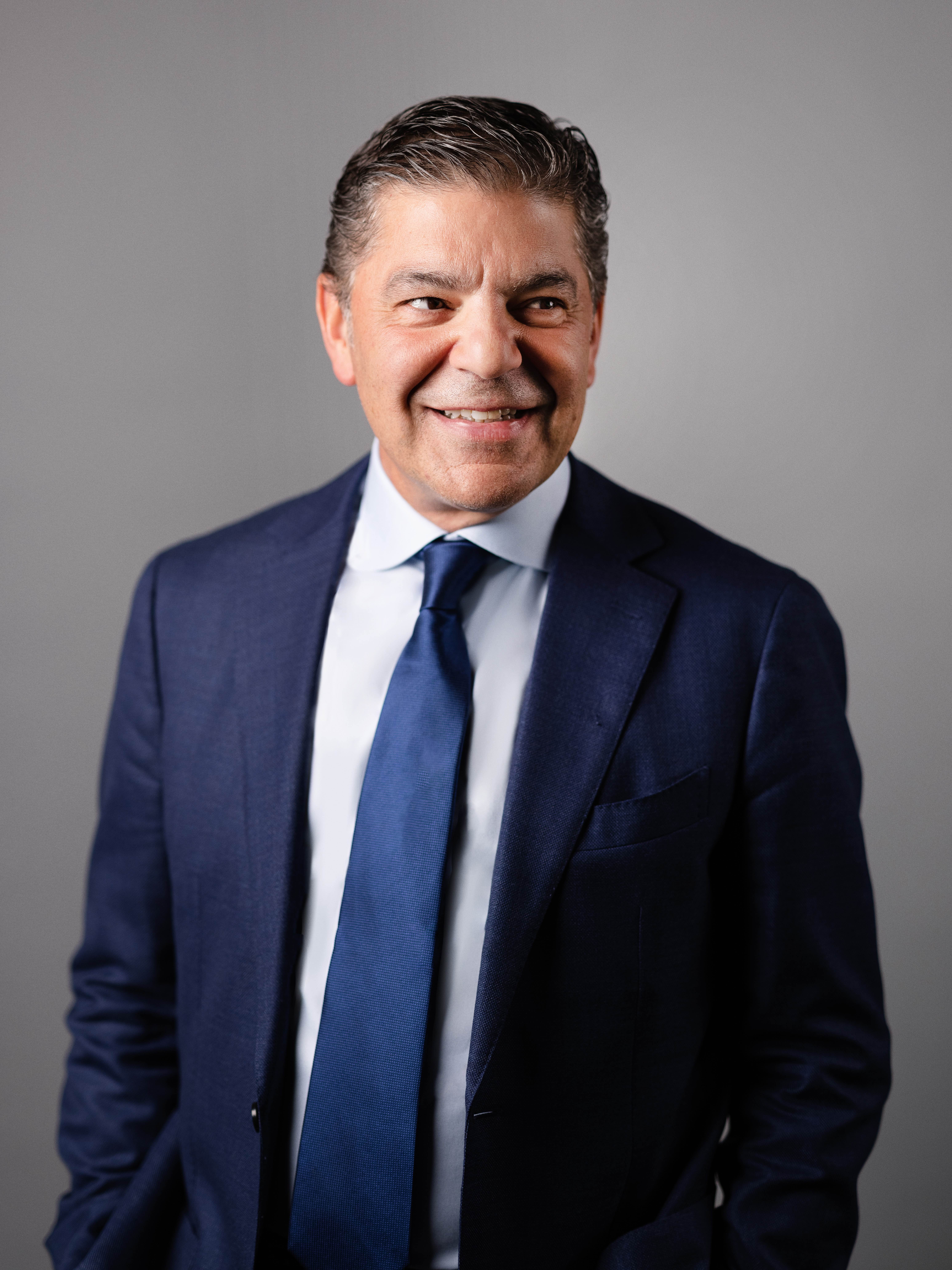 A smiling man with dark hair in a blue suit and tie against a grey background.