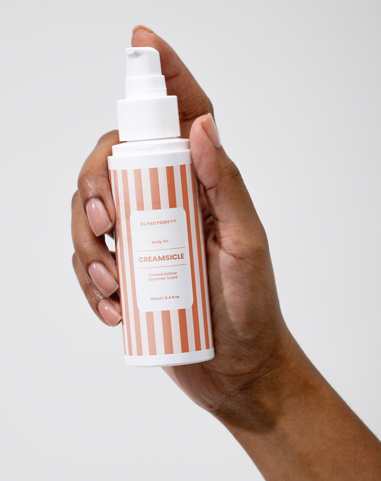 A hand holding a bottle of Olfactory NYC Creamsicle body oil against a plain background.
