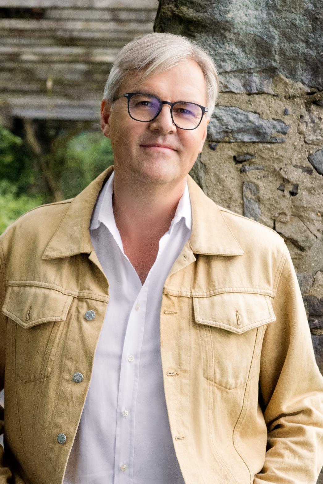 A man with graying hair and glasses, wearing a tan jacket, stands next to a stone wall.