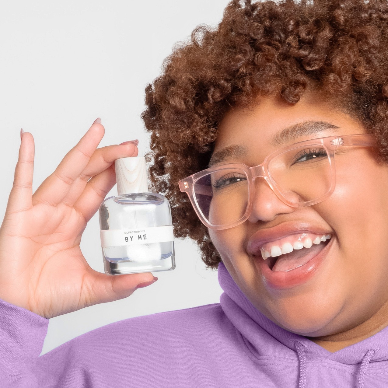 Person smiling and holding a perfume bottle labeled 'BY ME.'