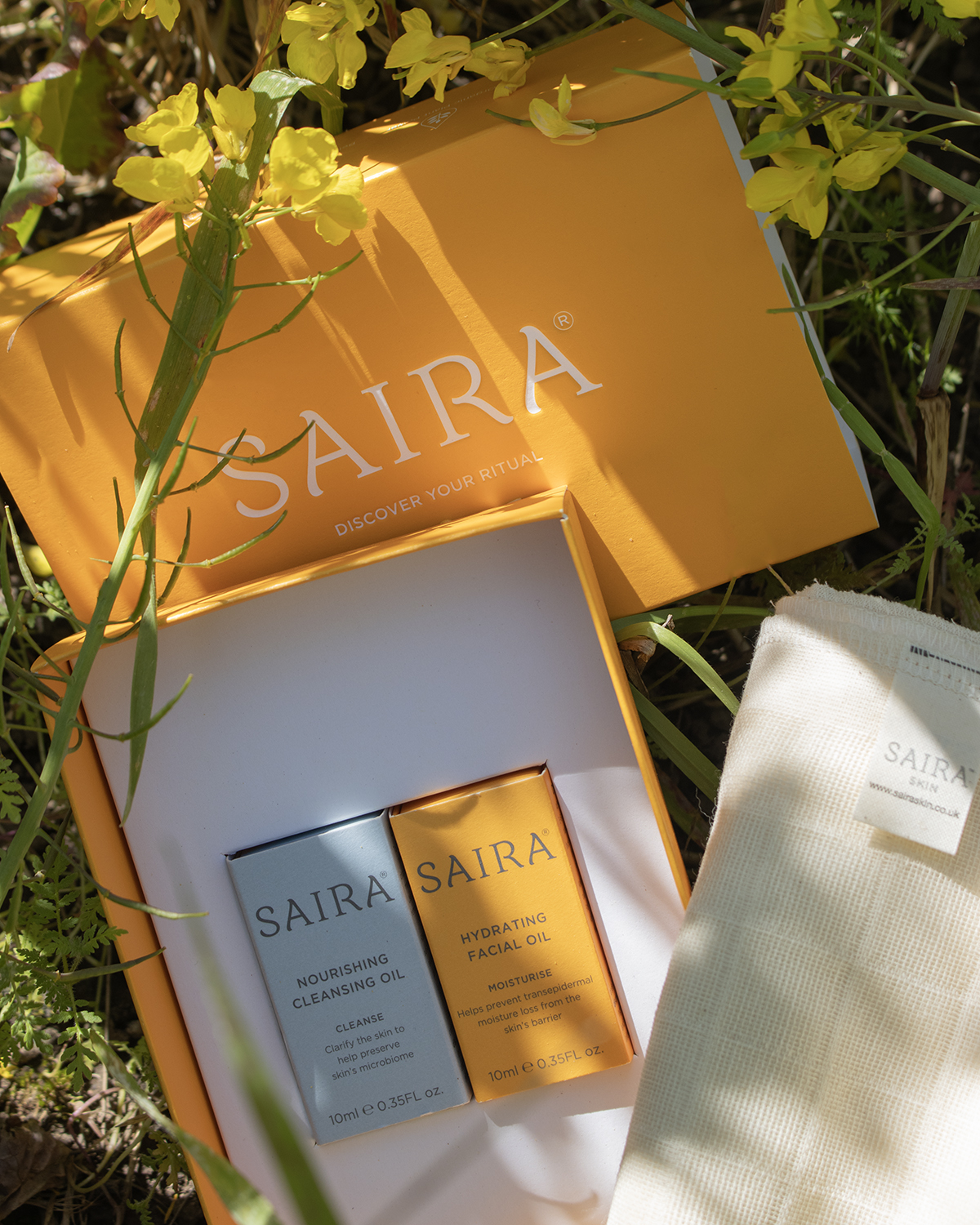 An open orange Saira skincare box containing two oils and a cloth, resting among yellow wildflowers.