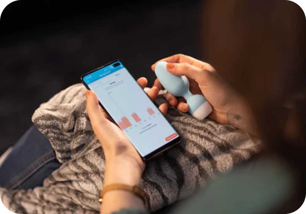A person sitting down, holding a smartphone with a graph on the screen and a small blue device.