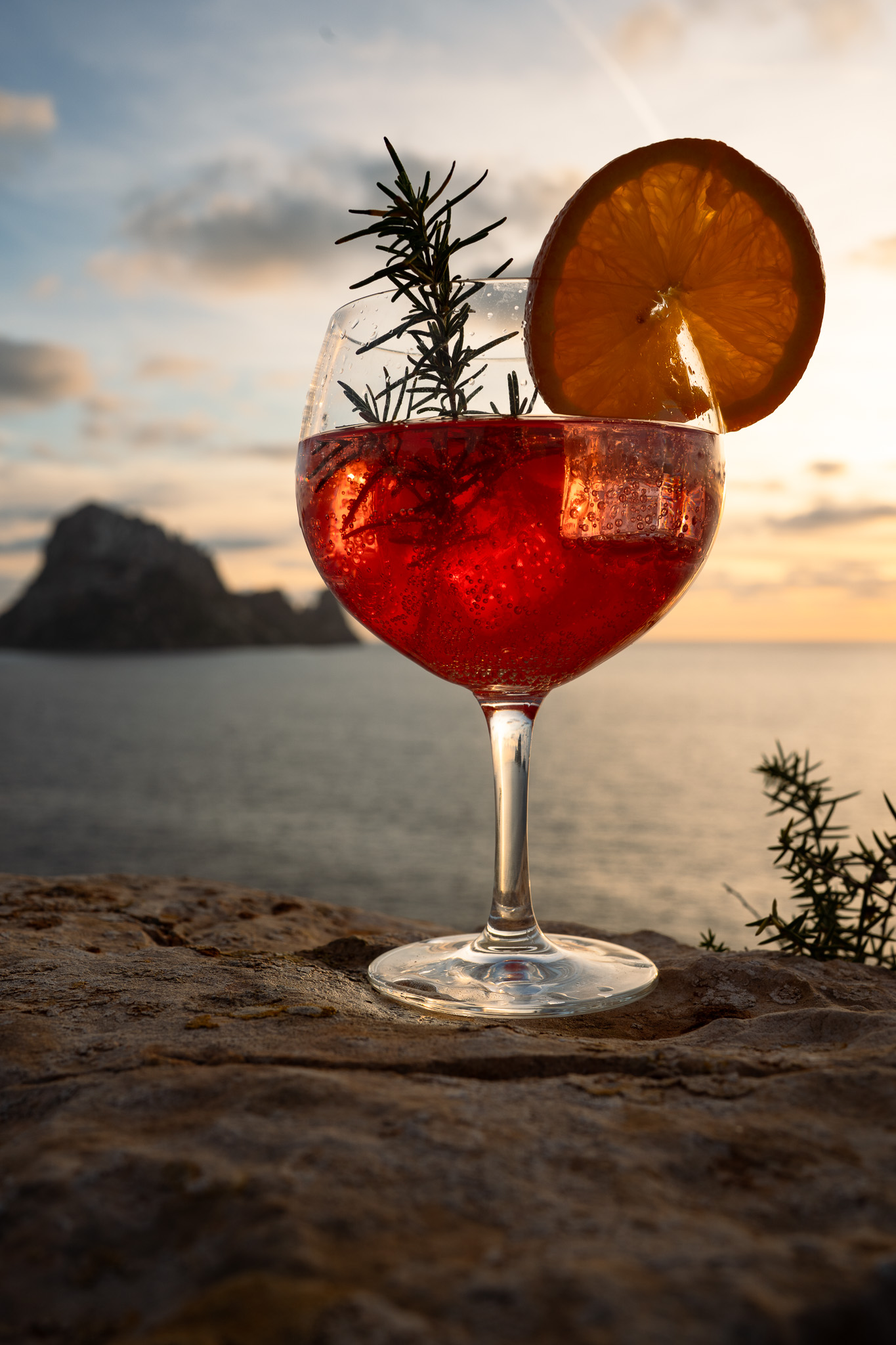 A person's arm, emerging from a pool, holds a red cocktail with an orange slice garnish.