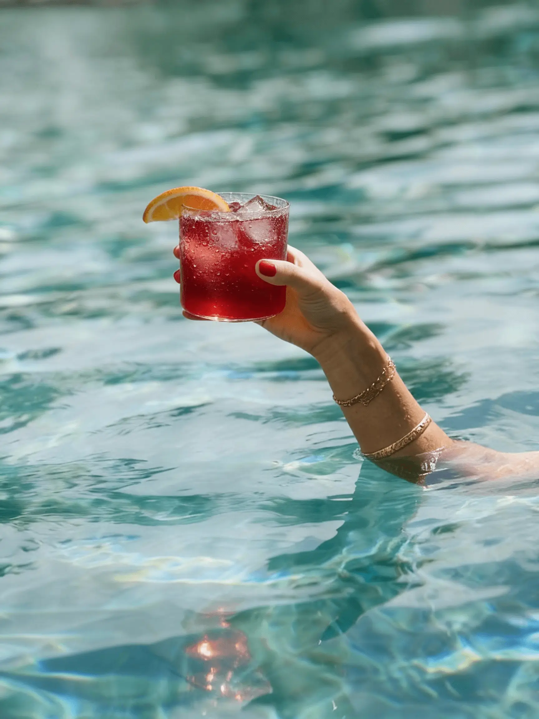 A person's arm, emerging from a pool, holds a red cocktail with an orange slice garnish.