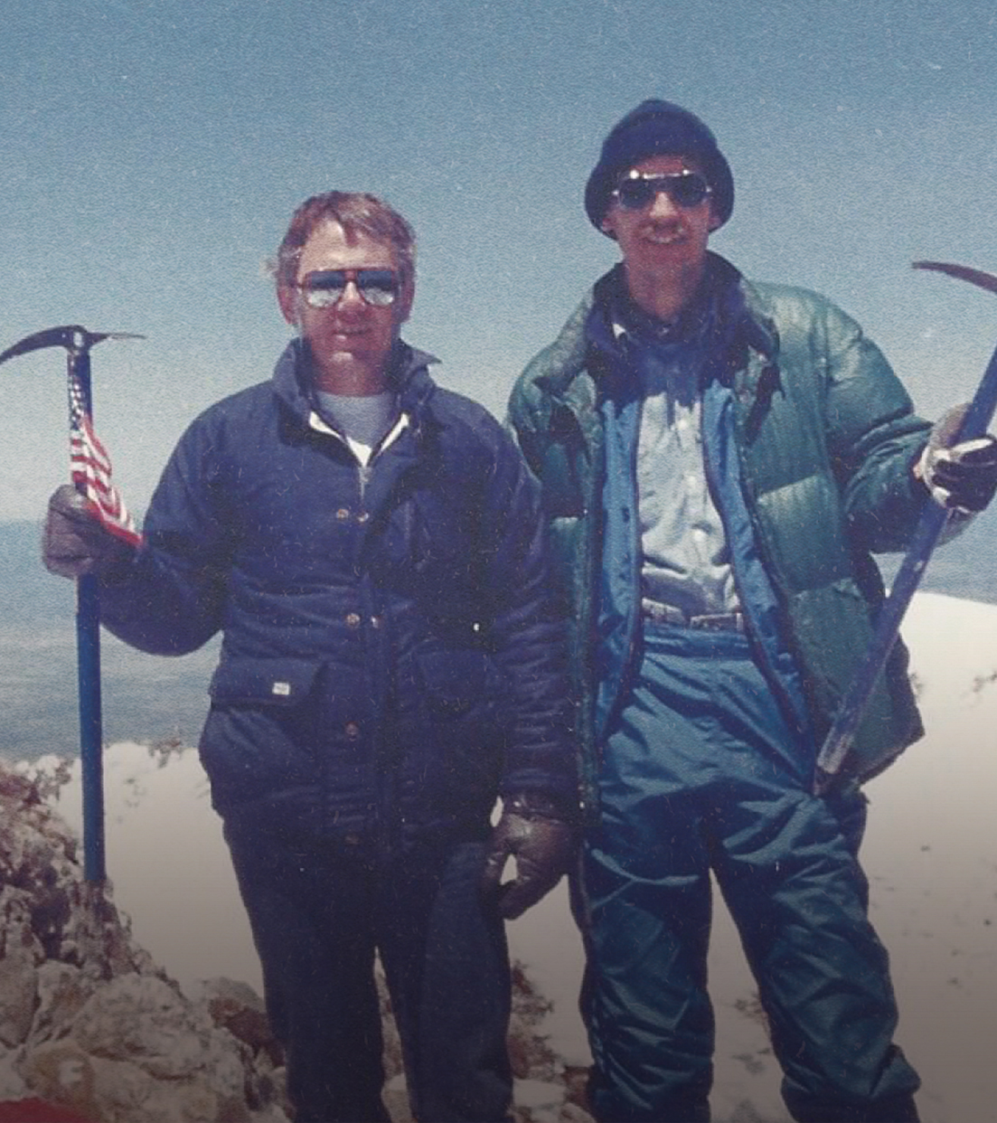 A vintage photograph of two men in winter jackets and sunglasses holding ice axes on a snowy mountaintop.