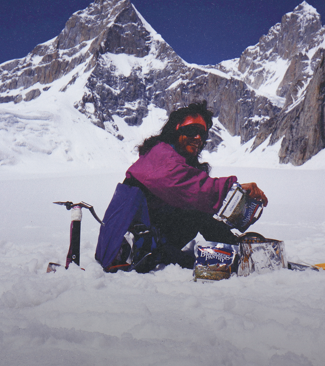 A mountaineer prepares food while sitting in the snow, with an ice axe nearby and mountains in the background.