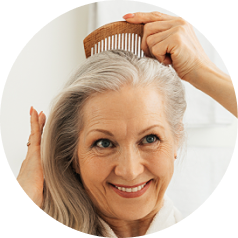 A smiling older woman with long grey hair combs her hair with a wooden comb.