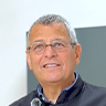 A person smiling, wearing glasses and a dark jacket, standing indoors.