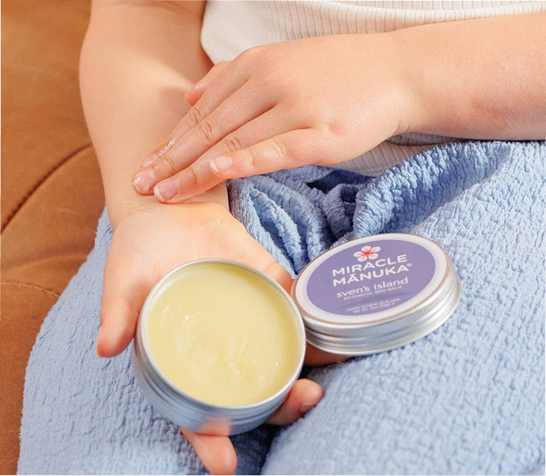 A person applies Miracle Mānuka skin balm from a small tin onto their inner wrist.