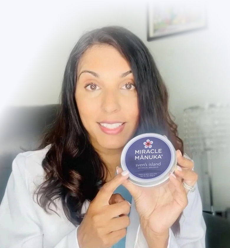A smiling woman in a white coat holds up a round tin of Miracle Mānuka botanical skin balm.