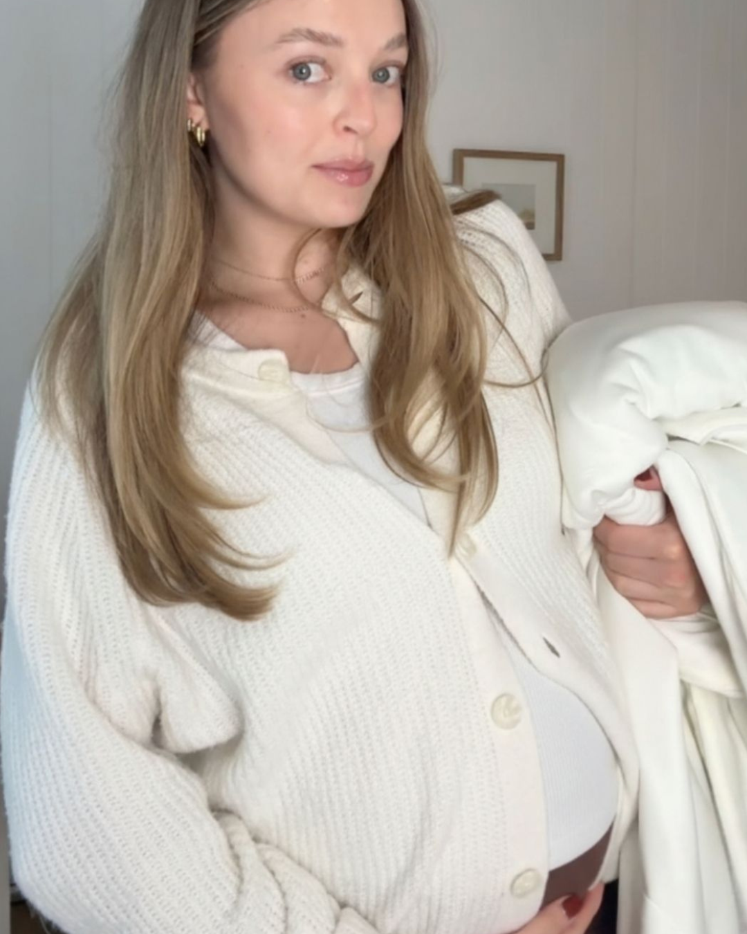 A pregnant woman in a white cardigan holds a white blanket and cradles her belly.