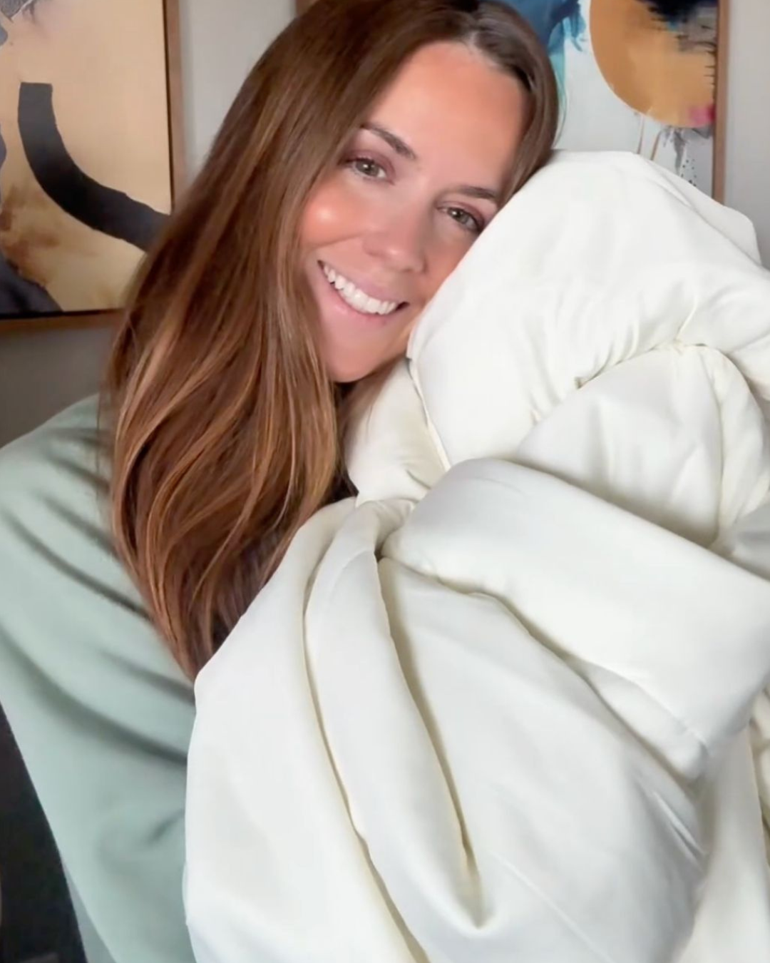 A smiling woman with long brown hair hugs a large, fluffy, off-white comforter indoors.