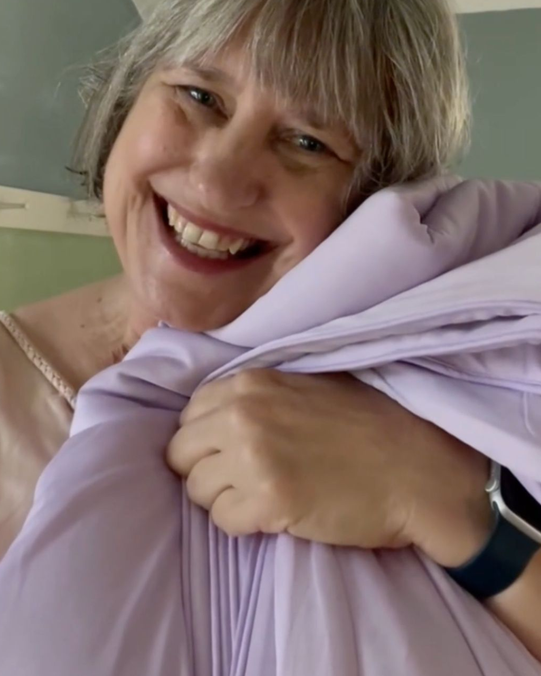 A close-up of a smiling woman with gray hair hugging a light purple pleated garment.