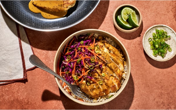 Bowl of rice with chicken, vegetables, and sauce, next to lime wedges and green onions.