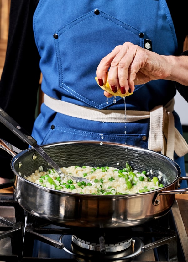 Person squeezing lemon over a pan of risotto with peas on a stove.
