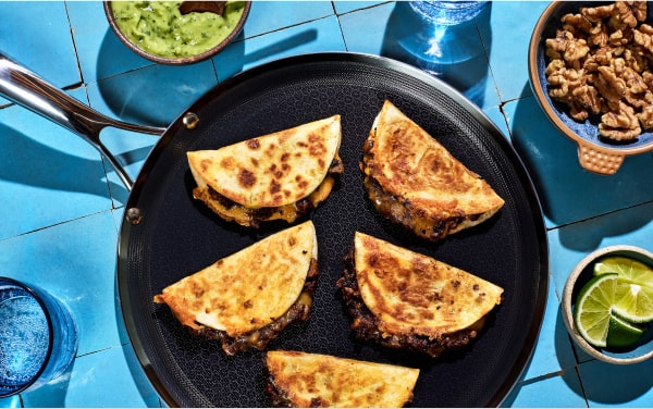 Five quesadillas on a skillet with guacamole, walnuts, and lime on a blue tiled surface.