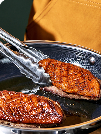 Two pieces of seared meat in a pan with tongs holding one piece.