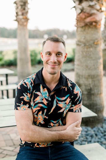 Person sitting outdoors by palm trees, wearing a floral shirt.