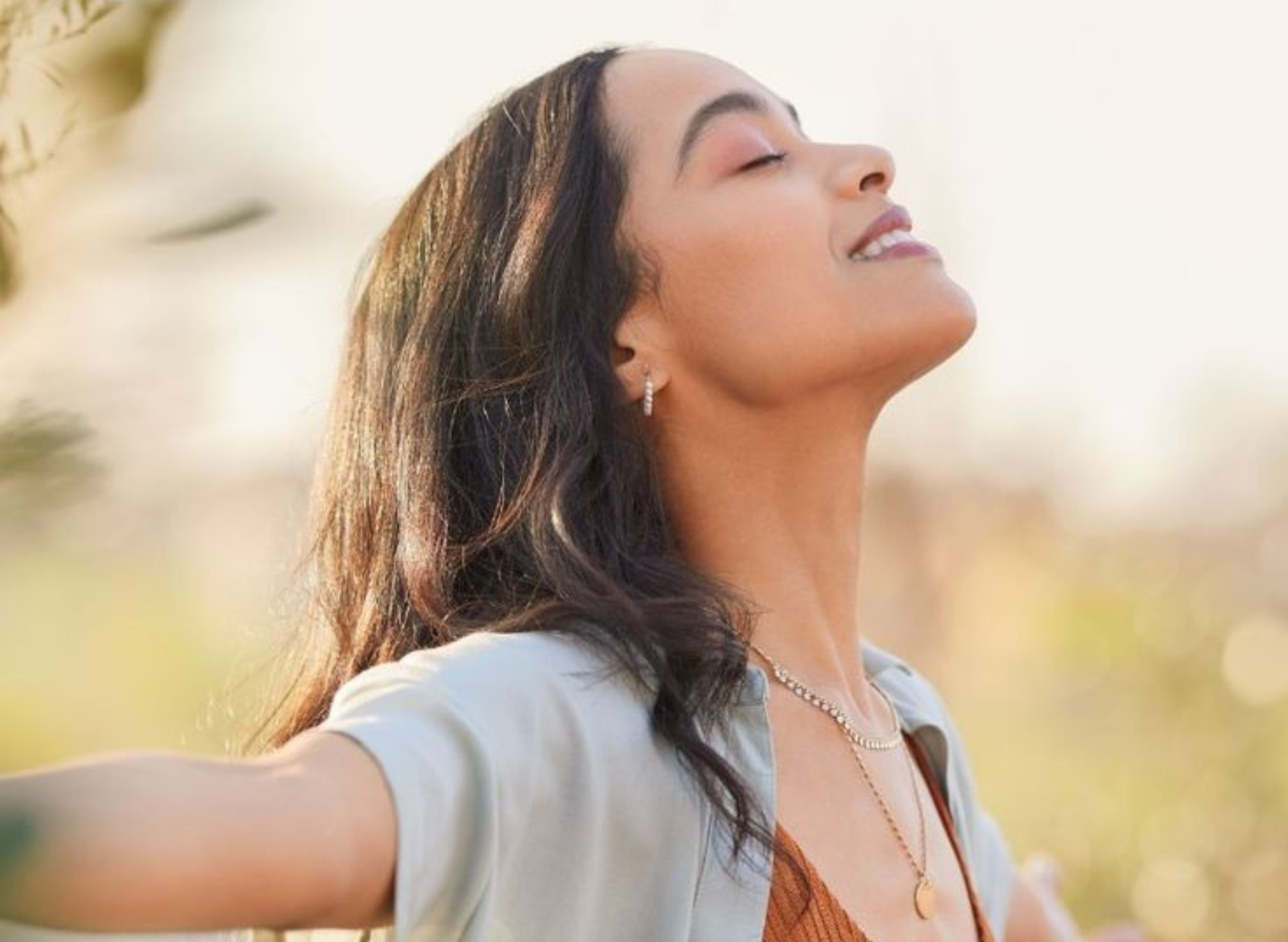 A woman with dark hair tilts her head back and smiles peacefully with her eyes closed outdoors.