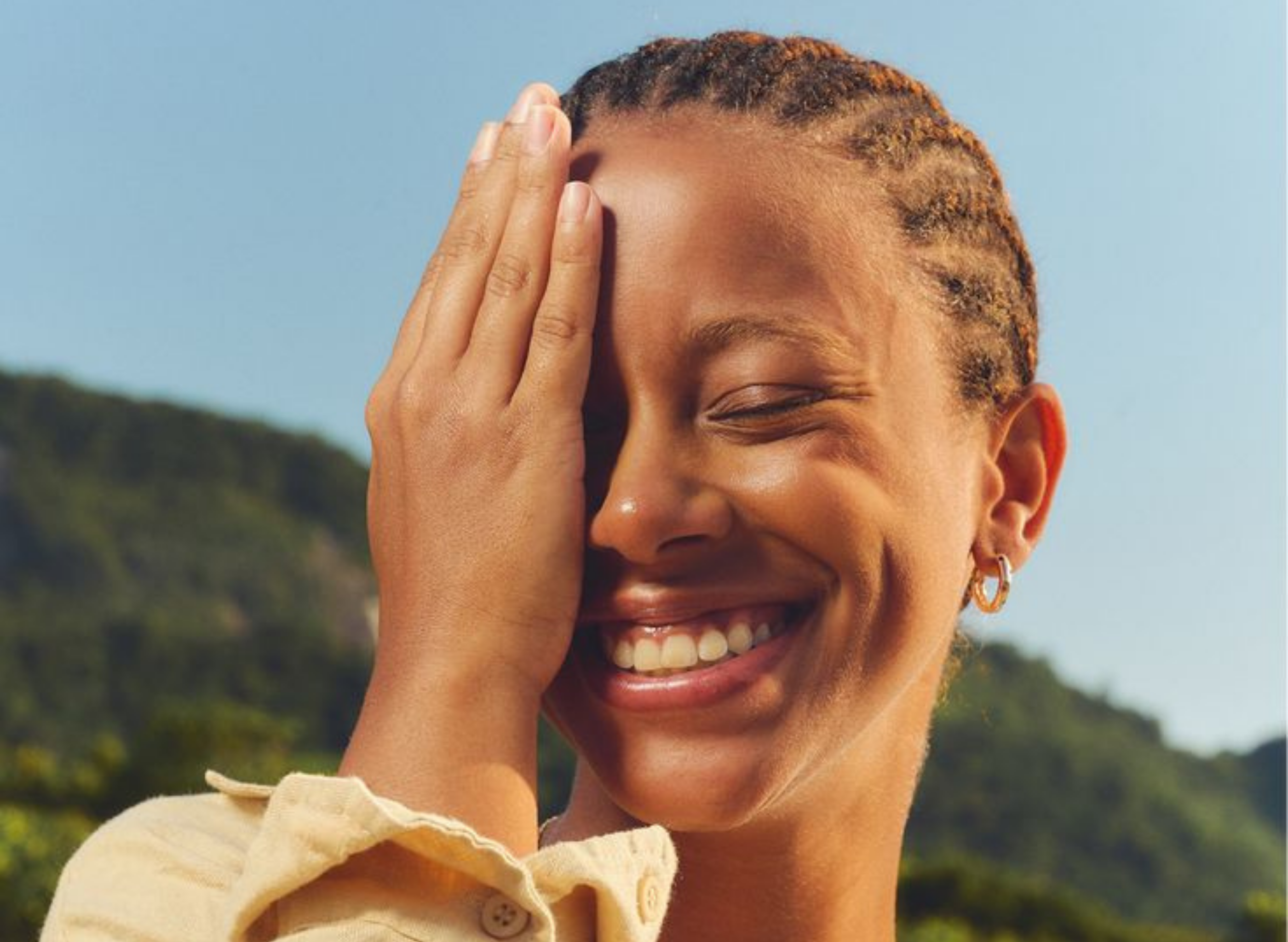 A smiling person with braided hair covers one eye with their hand while standing outdoors.