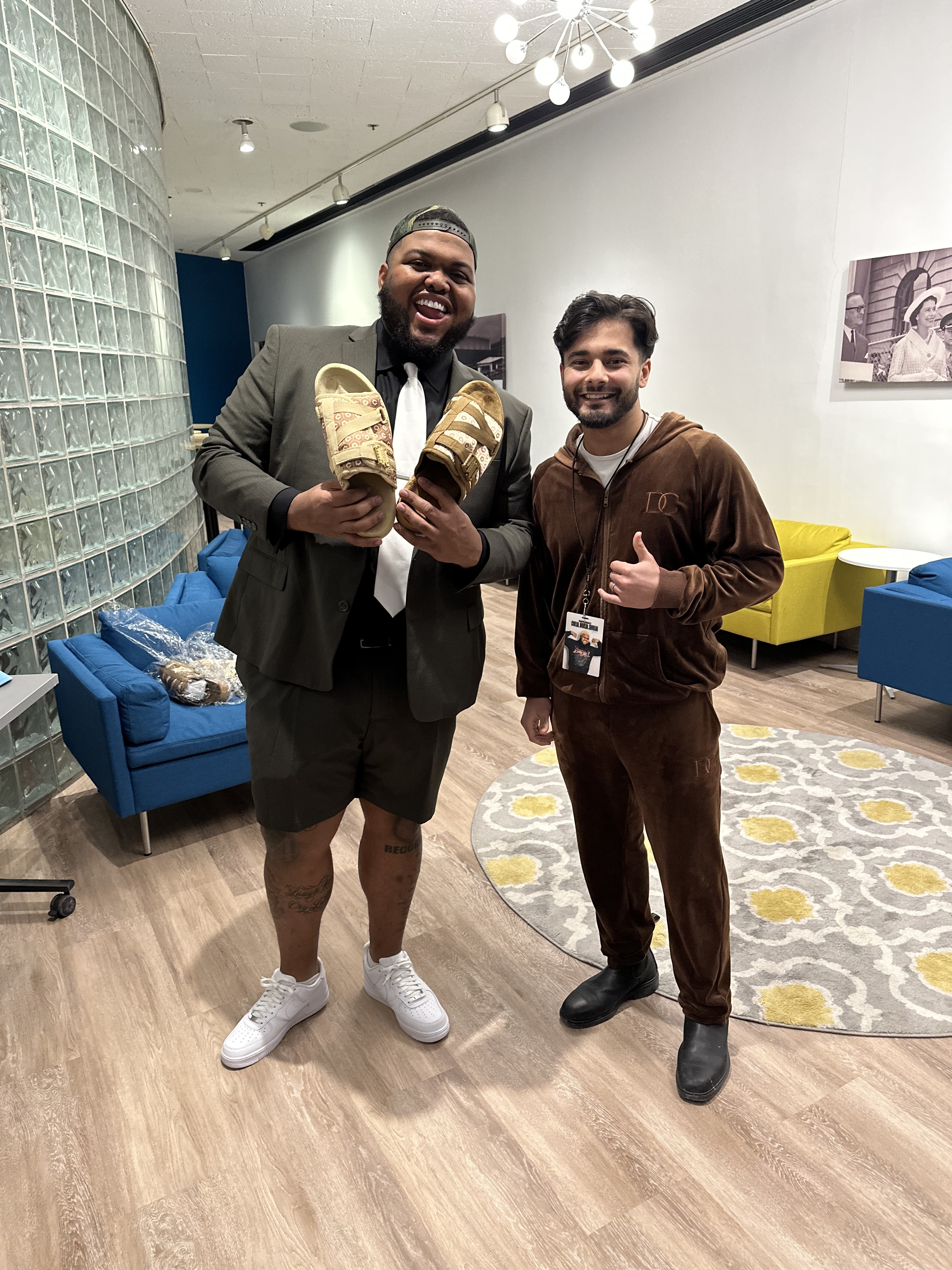 Two people standing indoors, one holding sandals, both smiling.