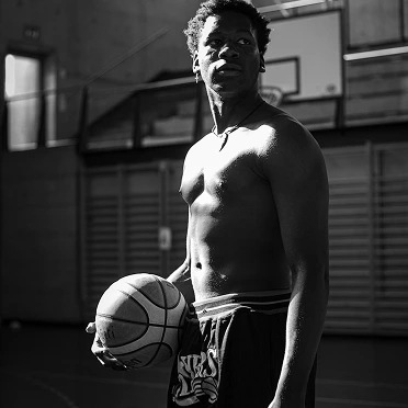 Shirtless man holding a basketball in an indoor gym, with dramatic lighting.
