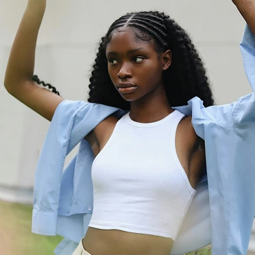 Person with braids and white top, blue shirt, arms raised outdoors.