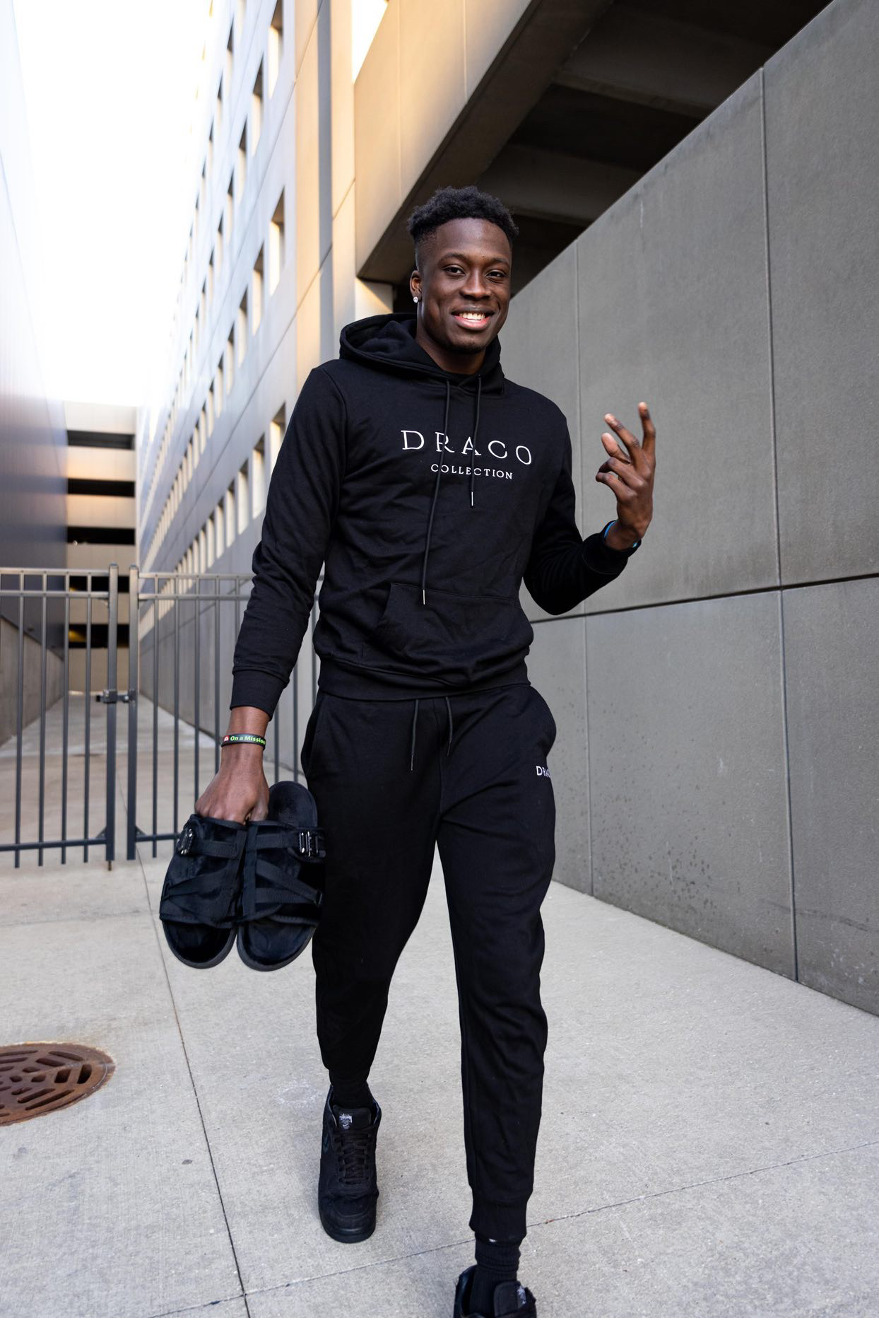 Person in black tracksuit smiling, holding sandals, walking outdoors.