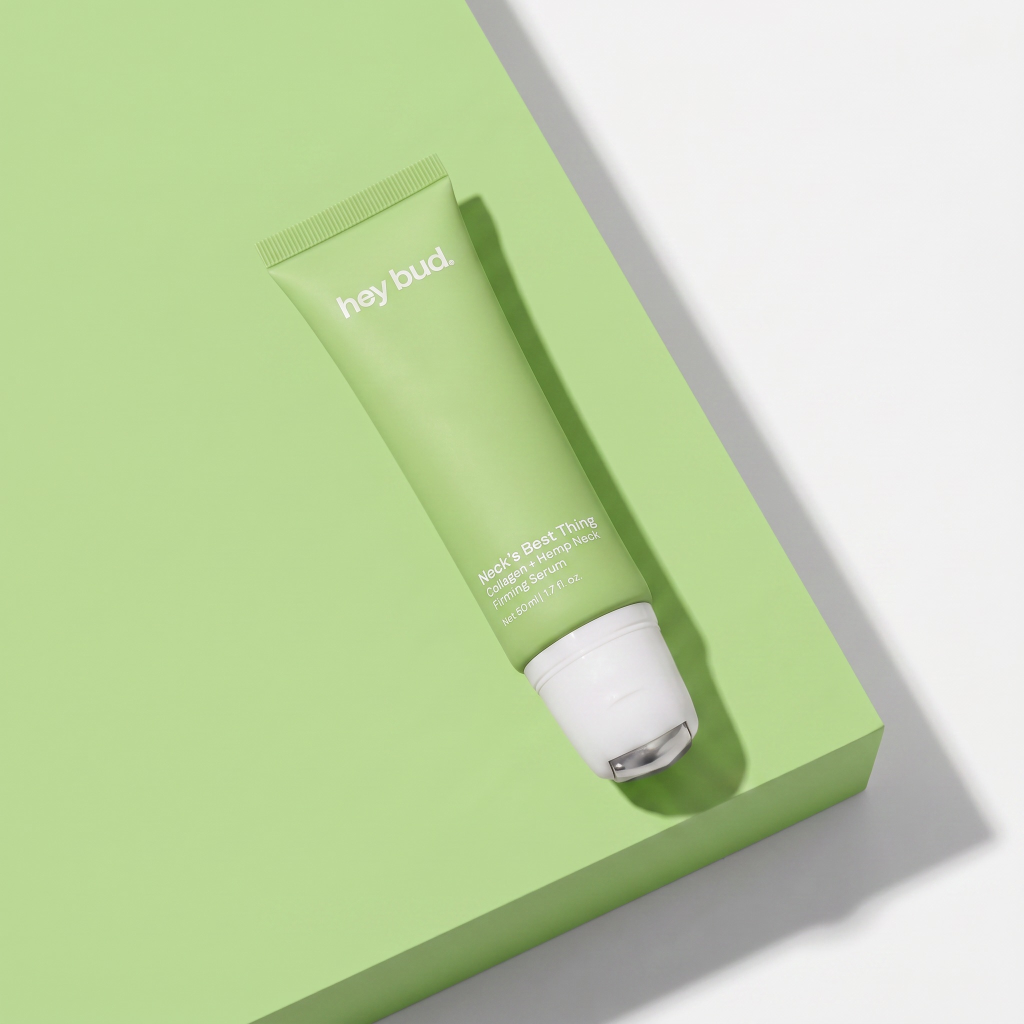 A light green tube of Hey Bud neck firming serum on a green and white geometric background.
