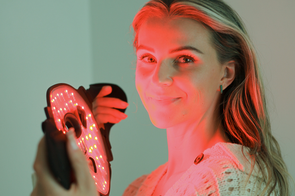 A smiling woman holds a glowing red LED light therapy mask next to her face.