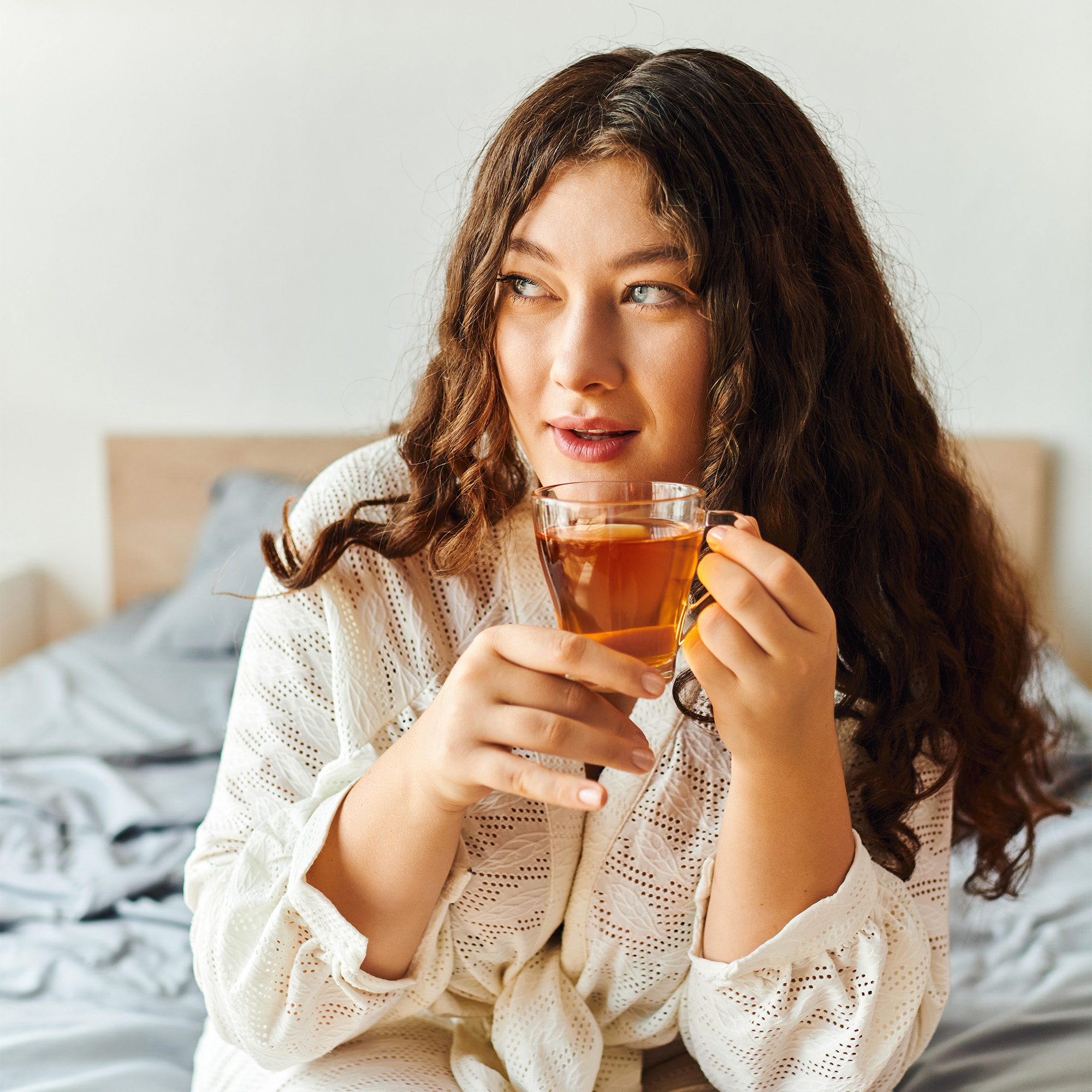 A woman with long curly hair sits in bed wearing a white robe and holding a glass of tea.
