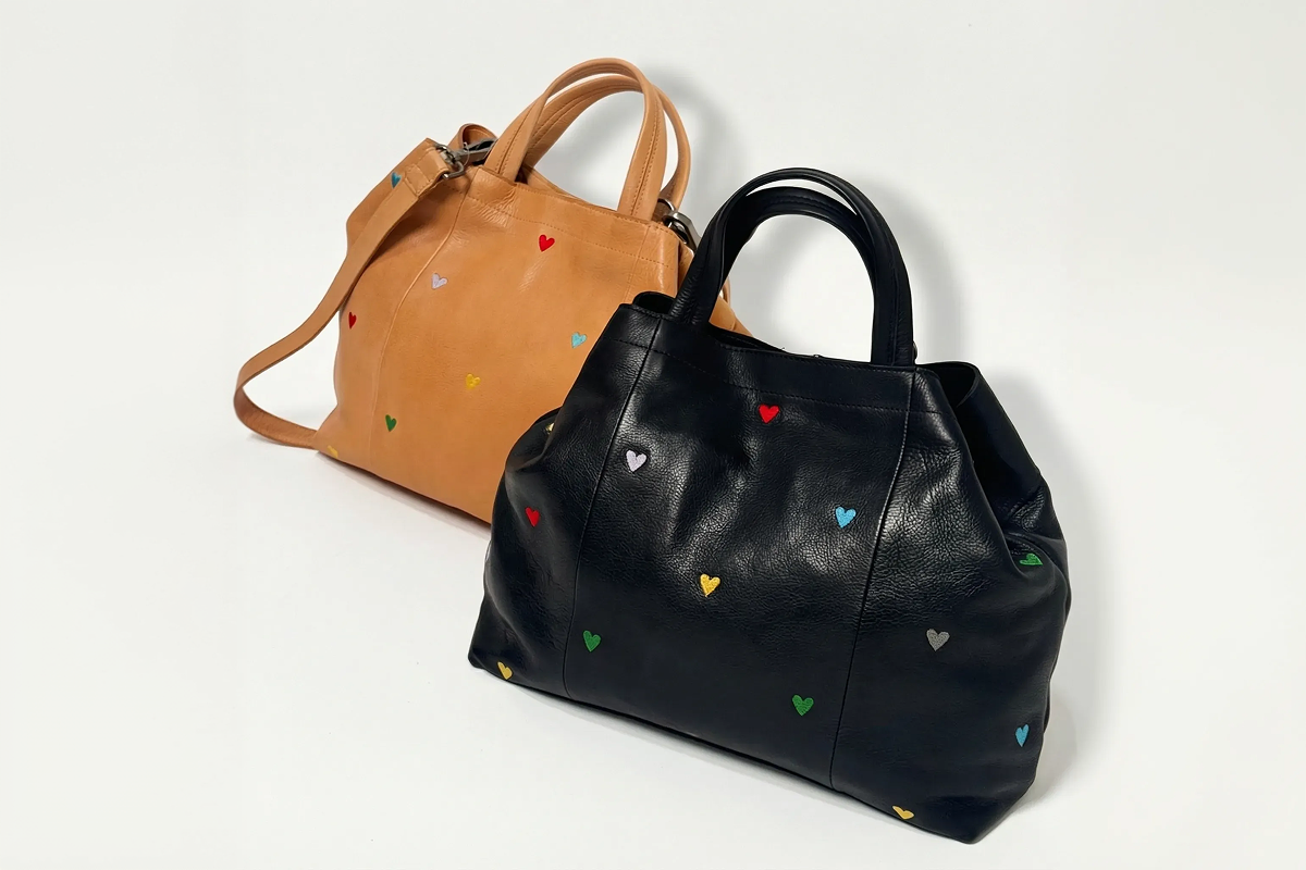 Two leather tote bags, one black and one tan, decorated with small, colorful embroidered hearts against a white background.