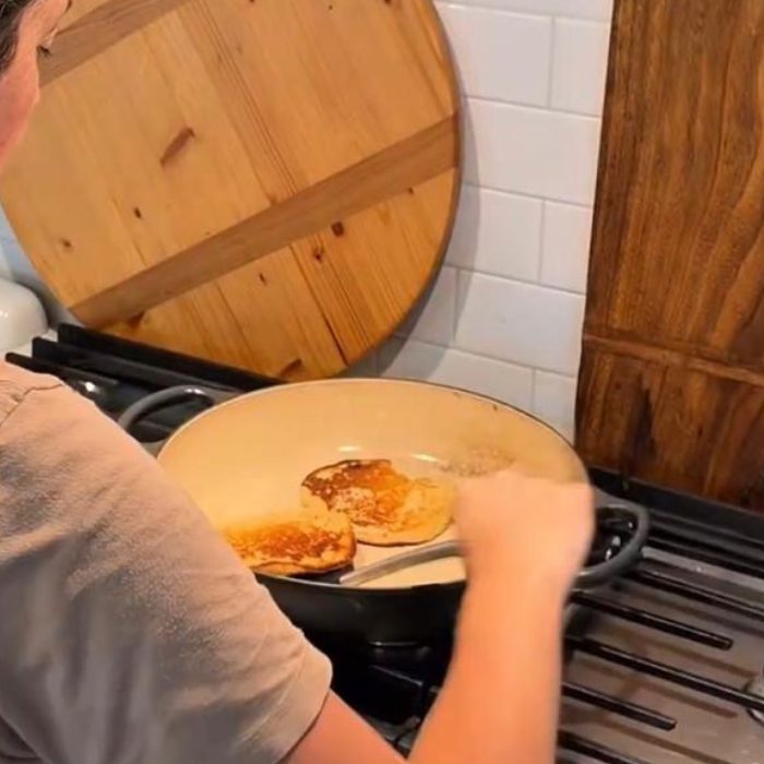 A person from behind cooking two pancakes in a pan on a stovetop.