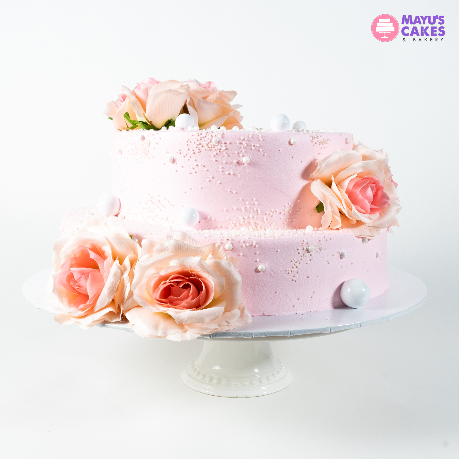 Two-tier pink cake with roses and decorative pearls on a white stand.