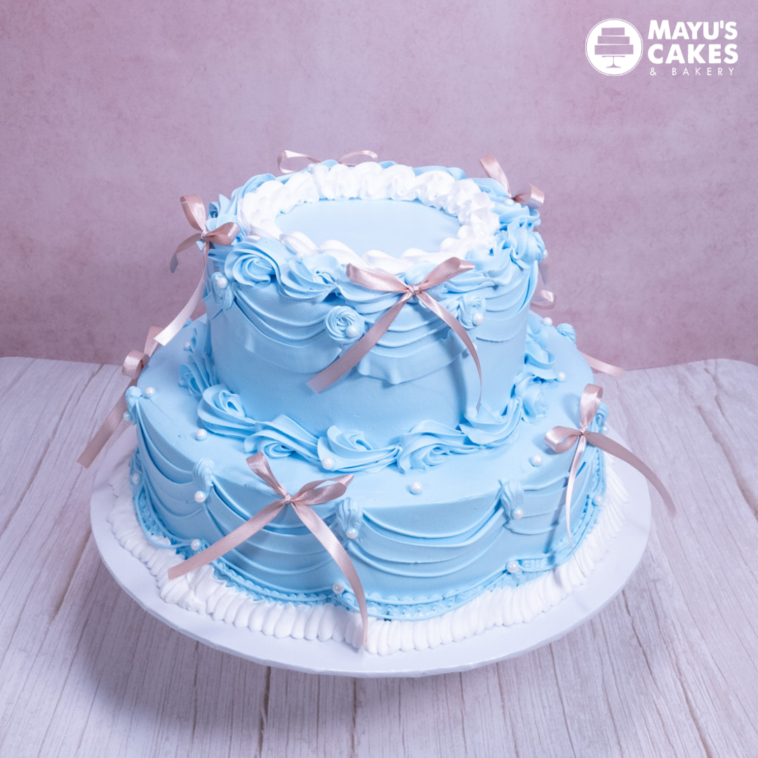 Two-tiered blue cake with pink ribbons and white decorations.