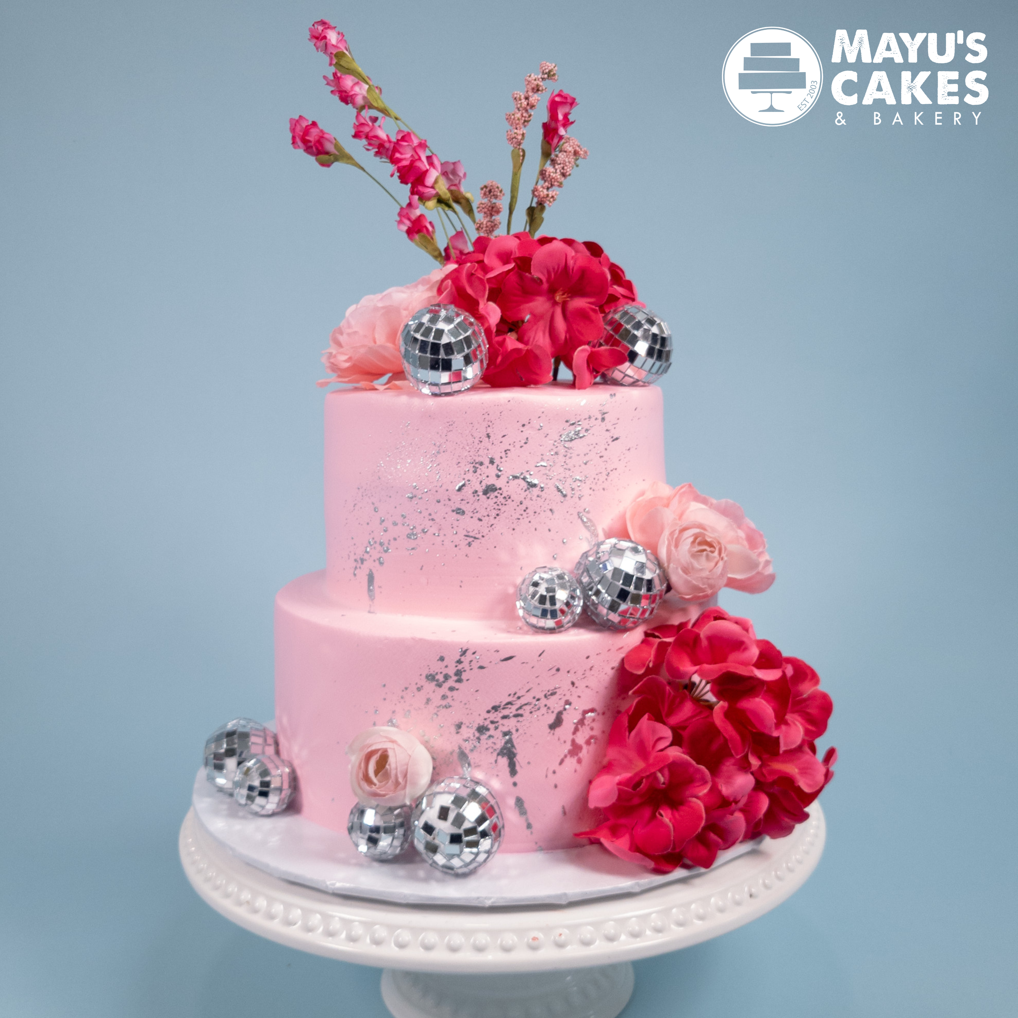 Two-tier pink cake with flowers and small disco balls on a white stand.