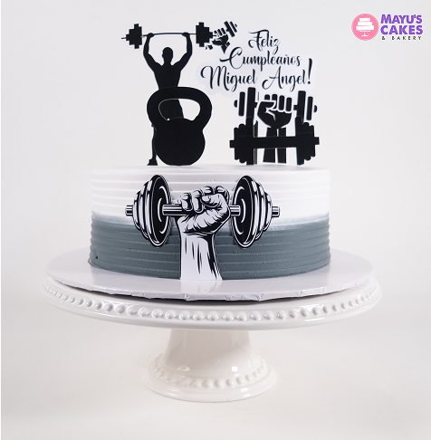 Gray and white cake with weightlifting decorations and 'Feliz Cumpleaños Miguel Angel' text.