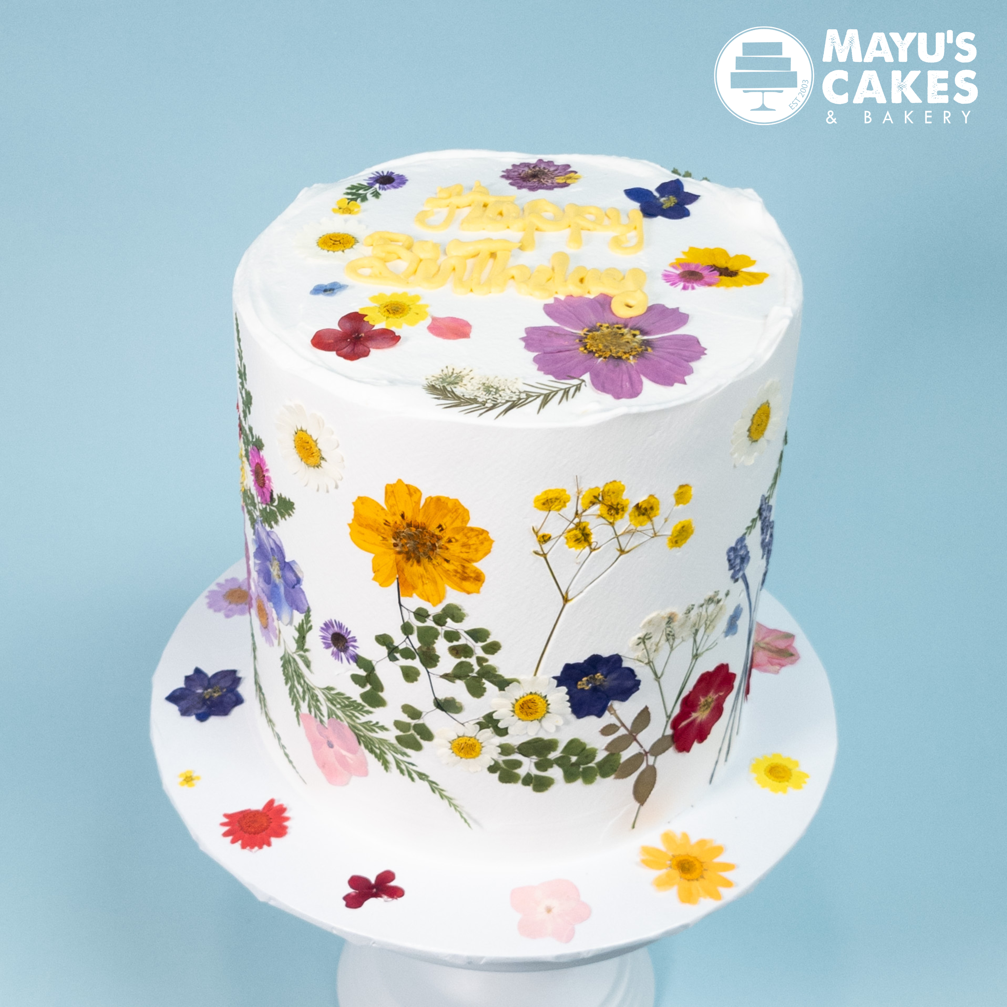 Tall white cake with colorful pressed flowers and 'Happy Birthday' text on top.