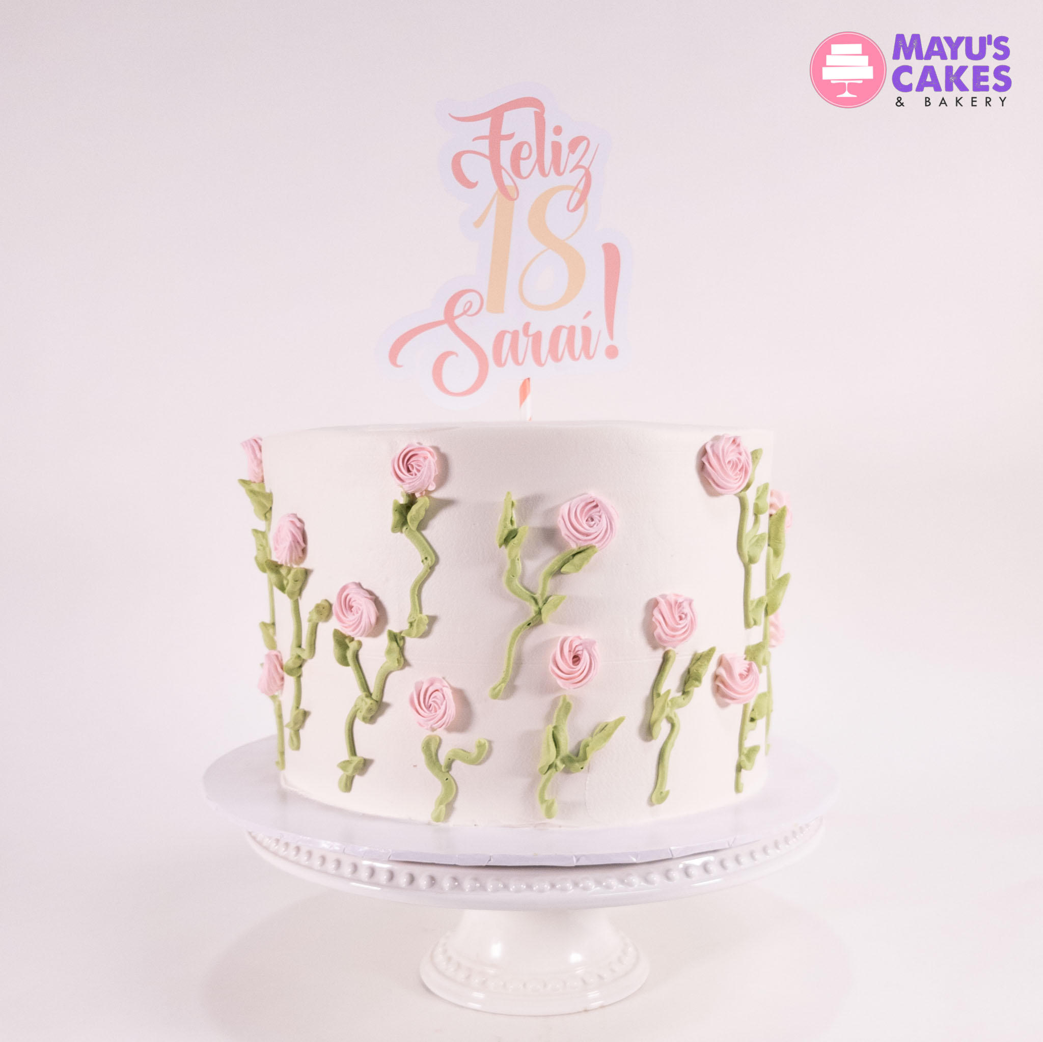 White cake with pink roses and 'Feliz 18 Sarai!' topper on a stand.