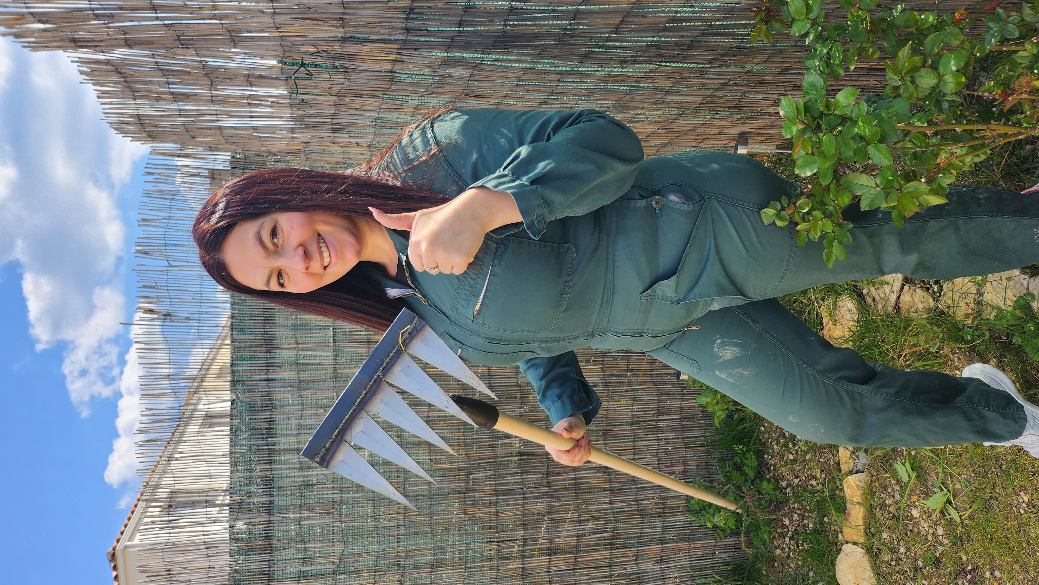 Woman in green overalls, holding garden tool, gives a thumbs-up outdoors.