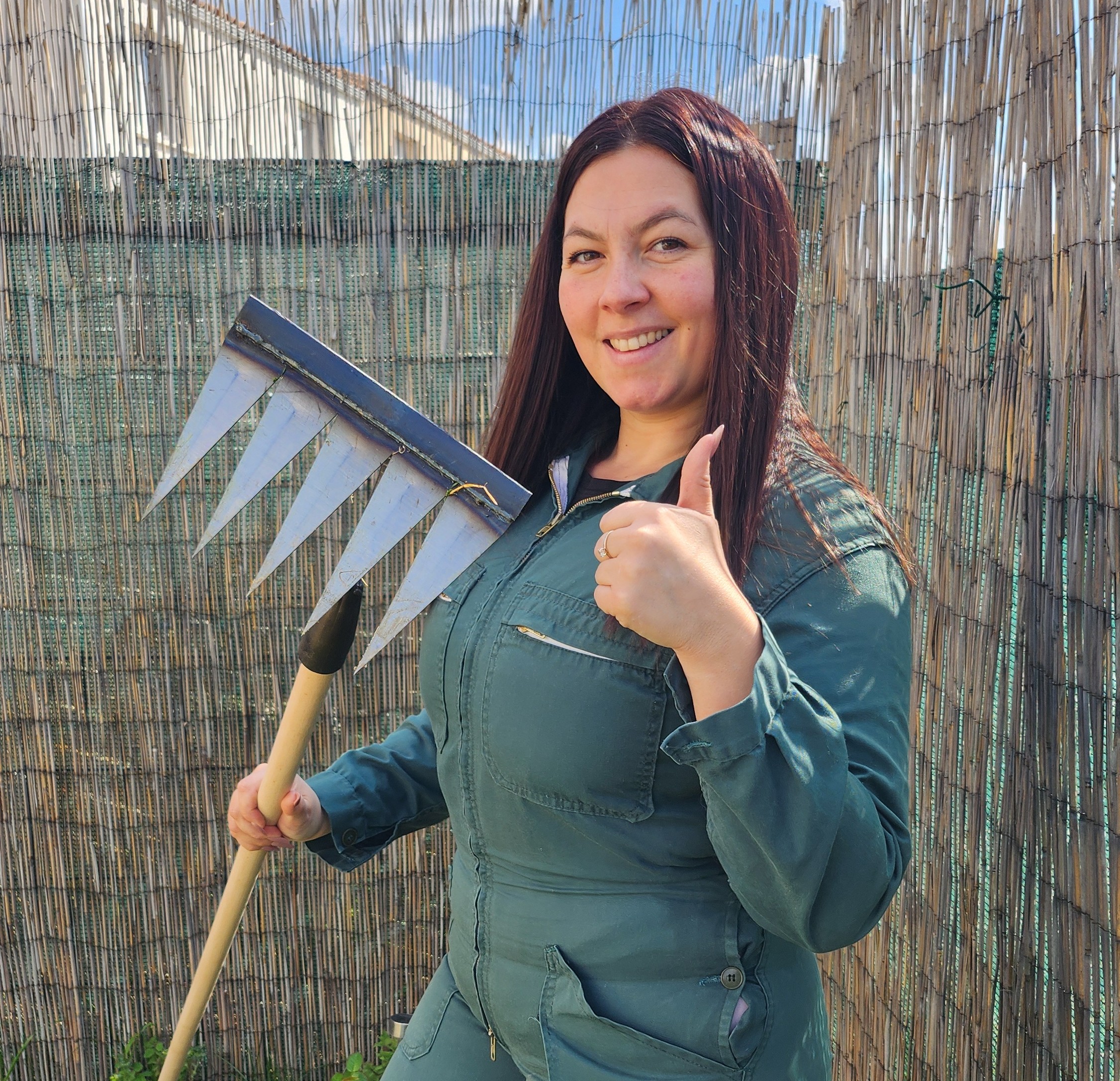 Person in green overalls holding rake, giving thumbs up in garden.