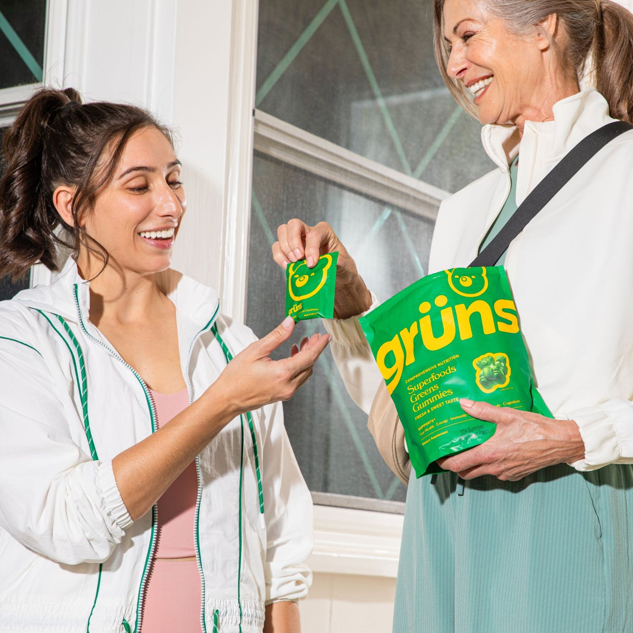 Two women smiling, holding green grüns gummy packages indoors.
