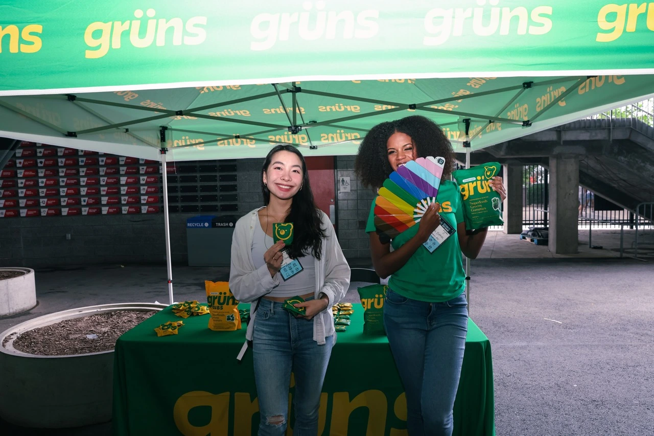 Two people under a green tent, holding colorful items and grüns products.