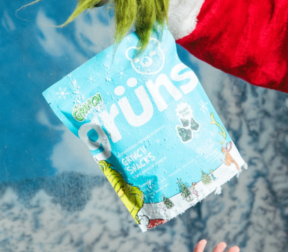 A Grinch-like hand holds a bag of Grinchy Snacks against a snowy, blue background.