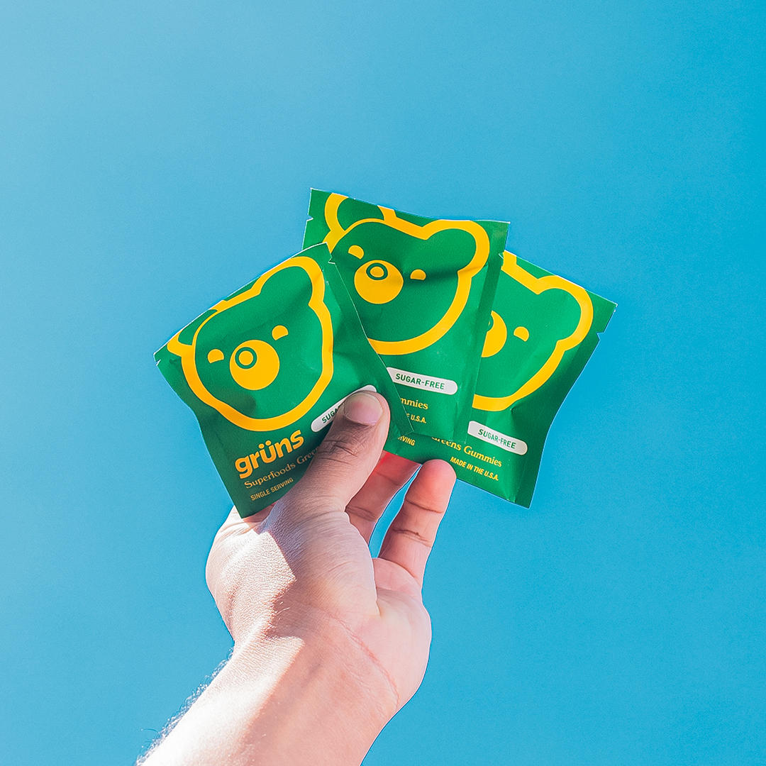 Hand holding three green gummy bear packets against a blue background.