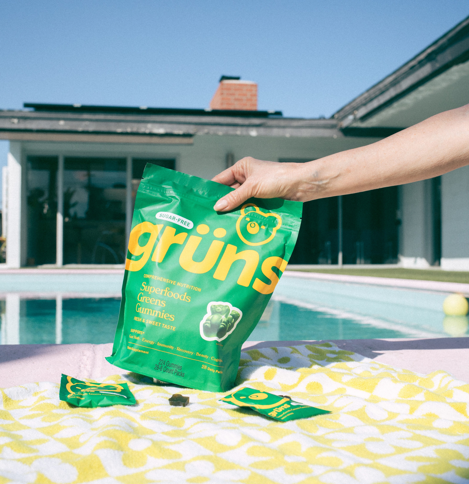 Hand holding grüns gummies pack by a poolside with a towel and smaller packs nearby.